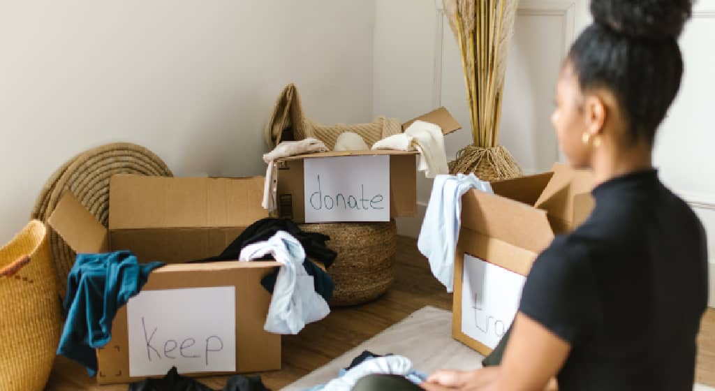 woman of color sitting crossed leg on the floor in front of three cardboard boxed labeled keep donate trash